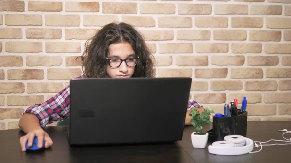 Δουλεύοντας σε ένα φορητό υπολογιστή, πληκτρολογώντας σε ένα πληκτρολόγιο, σύγχρονη έφηβος. Ένα εφηβικό αγόρι με σγουρά μαλλιά σε ένα καρό πουκάμισο χρησιμοποιεί ένα φορητό υπολογιστή σε ένα δωμάτιο στιλ σοφίτας. — Φωτογραφία Αρχείου