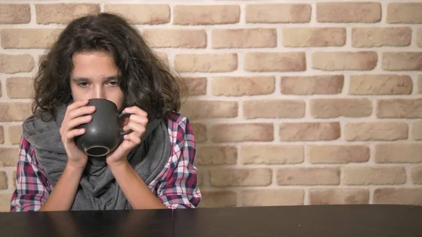 Het concept van seizoenskoorts. Zieke tiener jongen houdt een kopje warme drank. Ongelukkige tiener met krullend brunette haar, in een geruite overhemd en een grijze sjaal zit aan een tafel tegen een bakstenen muur — Stockfoto