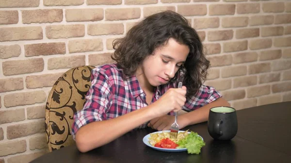 Adolescente faminto com apetite come farfalle, macarrão se curva na cozinha no estilo loft, contra uma parede de tijolo — Fotografia de Stock