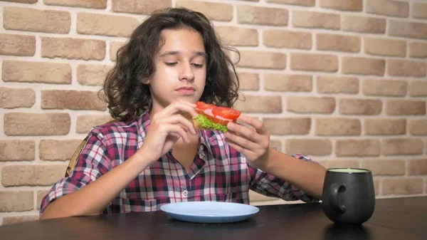 食欲を持つ空腹の十代の少年は、レンガの壁に対してロフトスタイルのキッチンで新鮮なレタスとトマトとサンドイッチを食べます — ストック写真