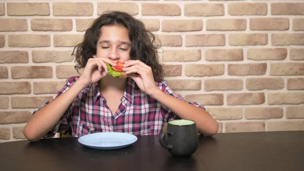 Adolescente faminto com um apetite come um sanduíche com alface fresca e tomates na cozinha estilo loft contra uma parede de tijolo — Fotografia de Stock