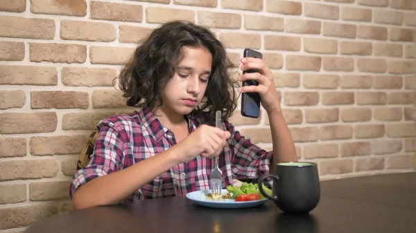 Hungrig tonåring pojke äter i köket och använder en smartphone. — Stockfoto