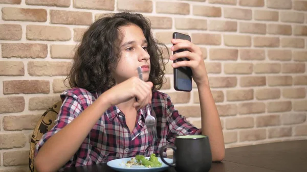 Hungrig tonåring pojke äter i köket och använder en smartphone. — Stockfoto