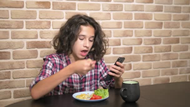 Fome adolescente menino come na cozinha e usa um smartphone . — Vídeo de Stock