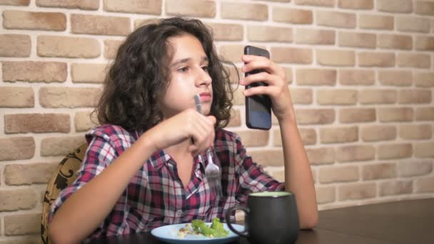 Голодный подросток ест на кухне и пользуется смартфоном . — стоковое видео