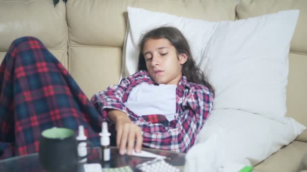 A teenage girl who has caught a cold at home on the couch, she covers herself with a blanket, measures the temperature. — Stock Video