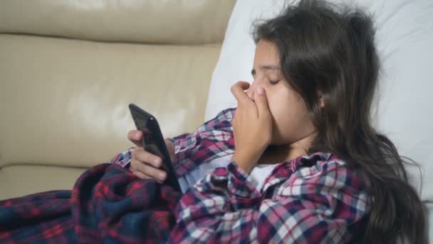 Ein krankes Mädchen hält ein Smartphone. Erkältetes Teenie-Mädchen deckt sich mit Wintergrippedecke ein. — Stockvideo