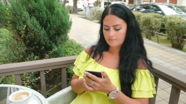 Jovem bela mulher morena sentada em um café ao ar livre, bebendo café e usando um telefone — Vídeo de Stock