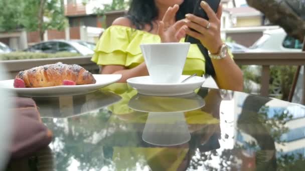 Joven hermosa morena sentada en una cafetería al aire libre, bebiendo café y usando un teléfono — Vídeos de Stock