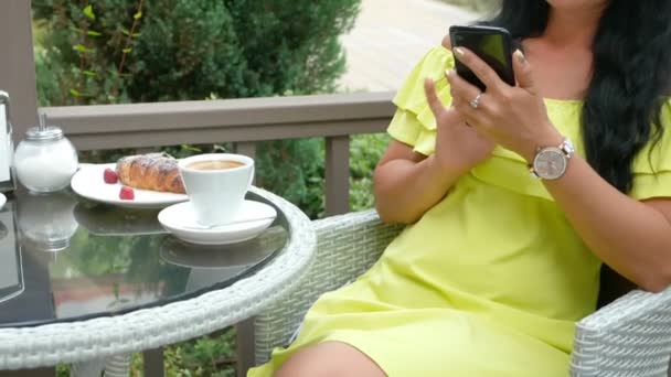 Νεαρή όμορφη μελαχρινή γυναίκα κάθεται σε ένα υπαίθριο καφέ, πίνοντας καφέ και χρησιμοποιώντας ένα τηλέφωνο — Αρχείο Βίντεο