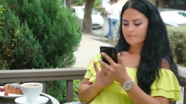genç güzel esmer kadın bir açık hava kafede oturan, kahve içme ve bir telefon kullanarak