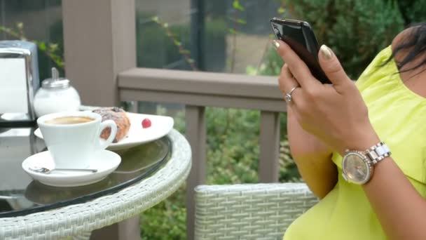 Joven hermosa morena sentada en una cafetería al aire libre, bebiendo café y usando un teléfono — Vídeos de Stock