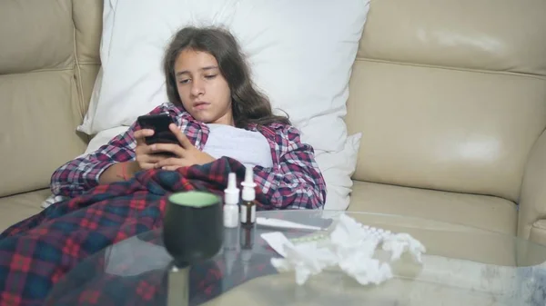 Menina doente segurando um smartphone. frio adolescente menina cobre com inverno gripe cobertor descansando em casa . — Fotografia de Stock