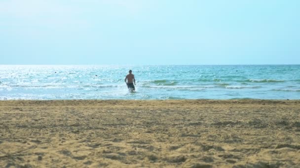 Молодой человек бегает по песчаному пляжу в море — стоковое видео