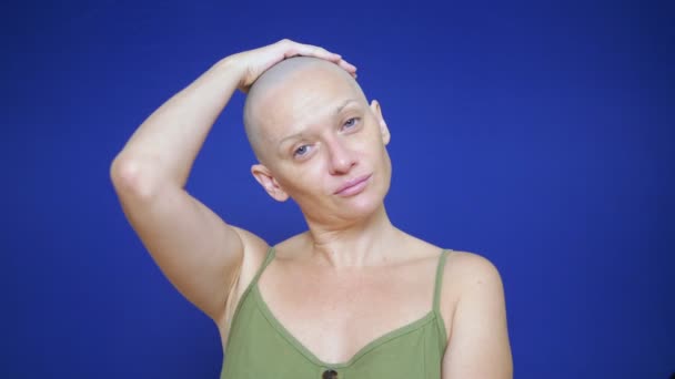 Лисий виснажена жінка на синьому фоні дивиться на камеру. вона почувається незручно через падіння волосся. концепція онкології та наслідки хіміотерапії — стокове відео
