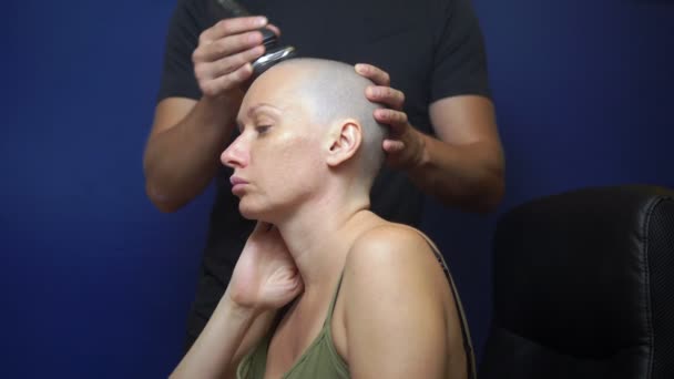 Man scheert een kale vrouw met een elektrisch scheermesje op een blauwe achtergrond. chemotherapie effecten concept. — Stockvideo