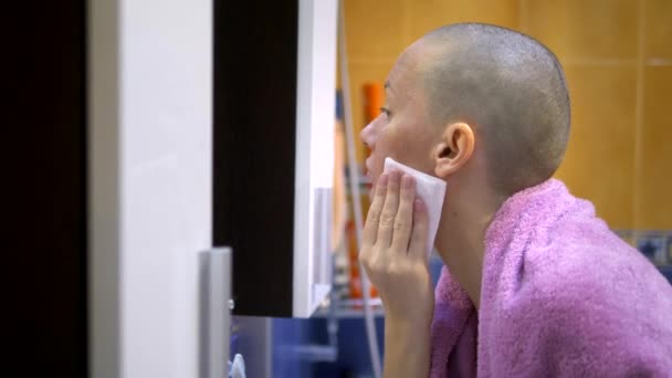 Stylish bald woman wipes her face with a napkin while looking in the mirror in the bathroom. — Stock Video