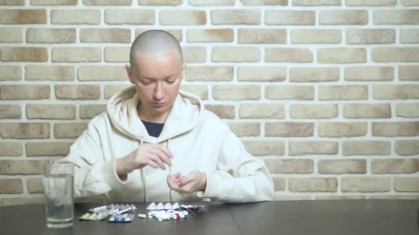 Uma jovem careca olha tristemente para as pílulas enquanto se senta em uma mesa contra uma parede de tijolos. espaço de cópia — Vídeo de Stock