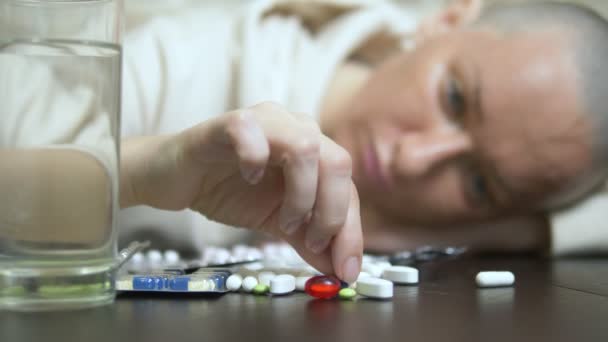 Uma jovem careca olha tristemente para as pílulas enquanto se senta em uma mesa contra uma parede de tijolos. espaço de cópia — Vídeo de Stock