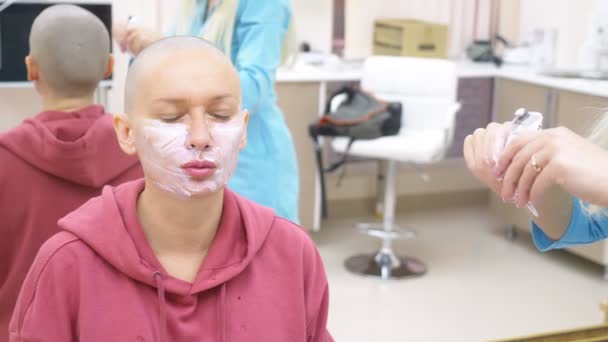 Snygg skallig kvinna på en kosmetolog utnämning. applicering av bedövnings kräm i ansiktet — Stockvideo