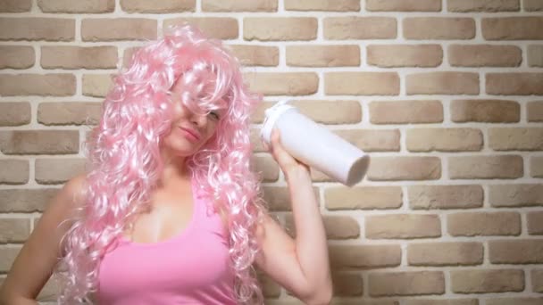 Funny cheerful girl with pink curly hair shakes a shaker against a brick wall background. copy space — Stock Video