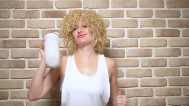 Счастливая веселая девушка с короткими белыми вьющимися волосами трясет шейкер на фоне кирпичной стены. копировальное пространство — стоковое видео