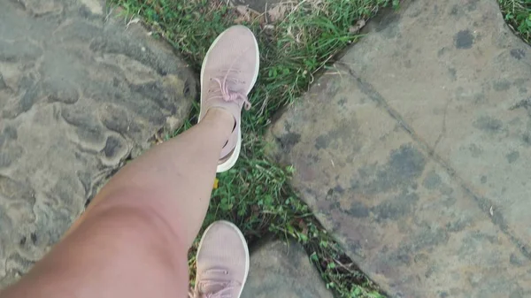 Pernas femininas em tênis rosa caminhar ao longo de um pavimento de paralelepípedos com grama e folhas caídas, vista em primeira pessoa — Fotografia de Stock