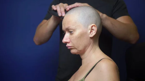 Hombre afeita a una mujer calva con una navaja eléctrica sobre un fondo azul. concepto de efectos de quimioterapia . — Foto de Stock