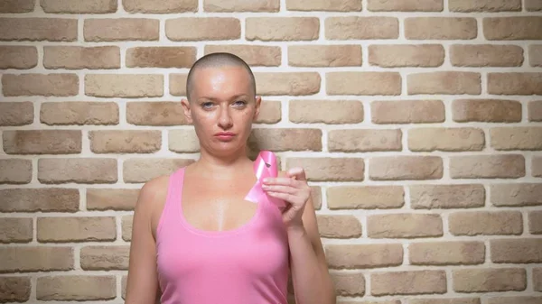 Mujer calva sobreviviente después del cáncer orgullosamente sostiene una cinta rosa en sus manos contra una pared de ladrillo . —  Fotos de Stock