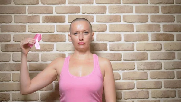 Bald woman Survivor after cancer proudly holds a pink ribbon in her hands against a brick wall. — Stock Photo, Image