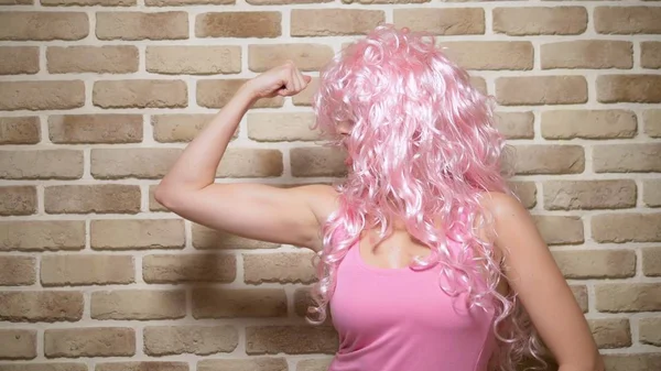 Menina louca com cabelo encaracolado rosa demonstra seu bíceps contra uma parede de tijolo. espaço de cópia. conceito de humor, aventuras de pessoas estranhas . — Fotografia de Stock