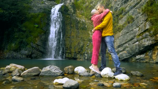 一对夫妇，一个年轻的男人和一个秃头的女孩在瀑布的背景下拥抱和亲吻。 旅行和探亲假概念。 复制空间 — 图库视频影像