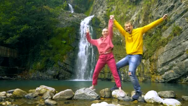 Um casal, um jovem e uma menina careca estão dançando com os braços levantados contra o fundo de uma cachoeira e olhando para a câmera. Viagens e conceito de férias em família. espaço de cópia — Vídeo de Stock