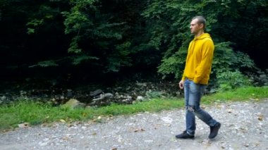 Sarı kazaklı ve kot pantolonlu genç bir adam parkta ya da ormanda yalnız yürür..