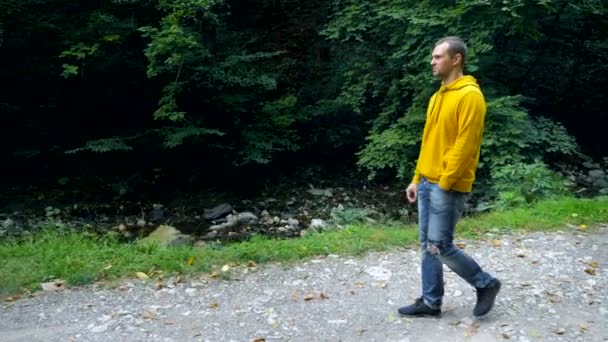 Een jongeman in een gele sweater en jeans loopt alleen in een park of bos. — Stockvideo
