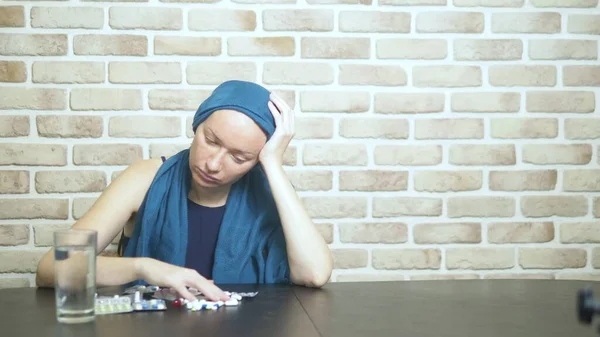 Een vrouw in een hoofddoek, een kankerpatiënt, raakt pillen, zittend aan een tafel tegen een bakstenen muur. ruimte kopiëren — Stockfoto