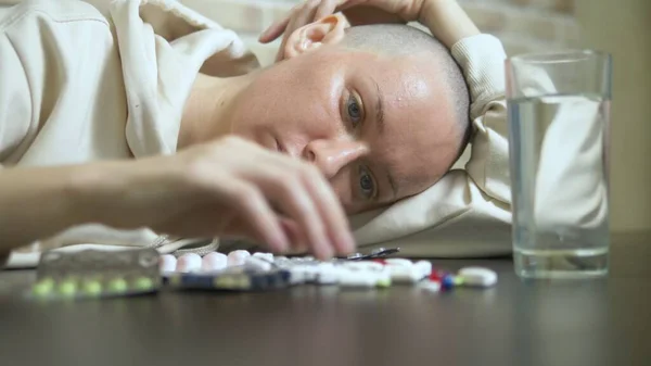 Primer plano. Una joven calva mira tristemente las pastillas mientras está sentada en una mesa contra una pared de ladrillo. espacio de copia — Foto de Stock