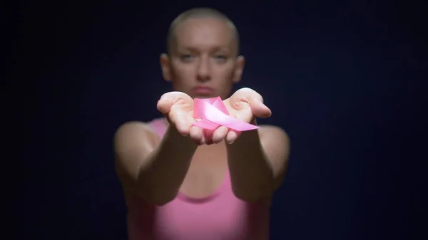 がんを生き延びたハゲの女性は、暗い背景にピンクのリボンを手に持っています。手とピンクのリボンに焦点を当てます。女性の顔がぼやけている. — ストック写真