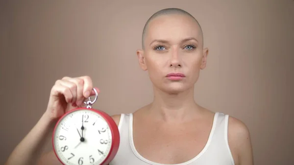 Bela mulher careca detém um despertador e uma fita rosa em suas mãos. fundo pastel suave . — Fotografia de Stock