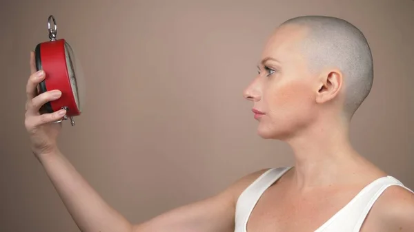 Retrato de mujer calva hermosa conmocionada con un reloj despertador sobre un fondo beige . — Foto de Stock