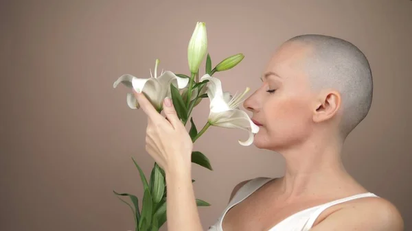 Beautiful smiling bald girl sniffs a white lily. beige background, copy space — Stock Photo, Image