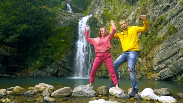 Um casal, um jovem e uma menina careca estão dançando com os braços levantados contra o fundo de uma cachoeira e olhando para a câmera. Viagens e conceito de férias em família. espaço de cópia — Vídeo de Stock