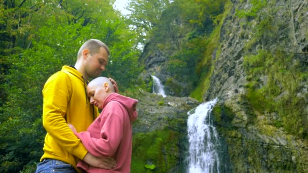 Pareja, un hombre joven y una chica calva abrazo y beso en el fondo de una cascada. Viajes y concepto de vacaciones familiares. espacio de copia — Vídeo de stock