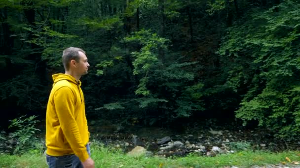 Egy fiatal férfi sárga pulóverben és farmerben egyedül sétál egy parkban vagy erdőben.. — Stock videók
