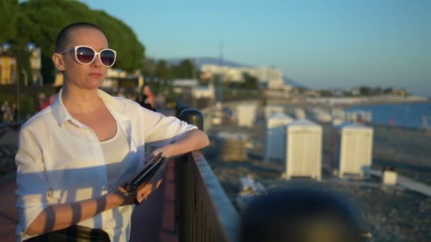 Femme chauve élégant dans les lunettes de soleil regarde le coucher du soleil. debout sur la promenade — Video