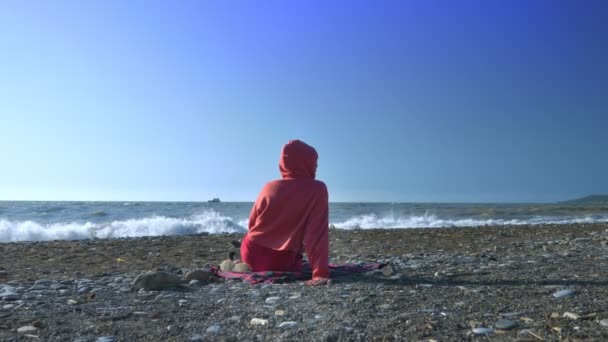 Vista posteriore. qualcuno in felpa rossa e cappuccio è seduto sulla spiaggia durante una tempesta e guardando il mare — Video Stock