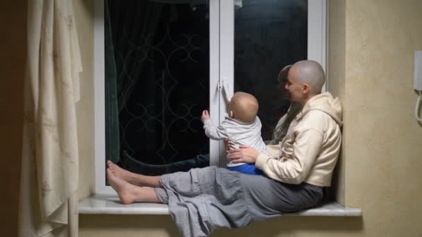 Frau mit Glatze und Baby sitzen abends zusammen am Fenster. — Stockvideo