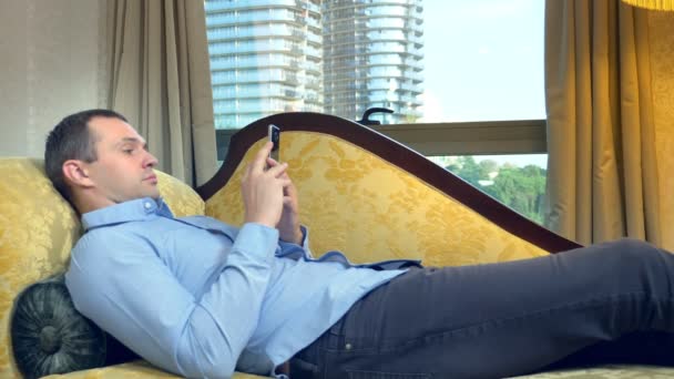 Empresário descansando em um sofá com um telefone contra uma janela panorâmica a partir da qual os arranha-céus são visíveis — Vídeo de Stock