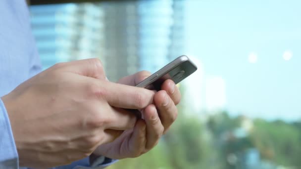 Nahaufnahme. Männliche Hände benutzen das Telefon vor dem Hintergrund eines Fensters, aus dem Wolkenkratzer zu sehen sind — Stockvideo