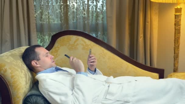 Hombre de negocios en un albornoz se relaja en un sofá con un teléfono y fuma un vapor en la noche en el fondo de una ventana — Vídeos de Stock